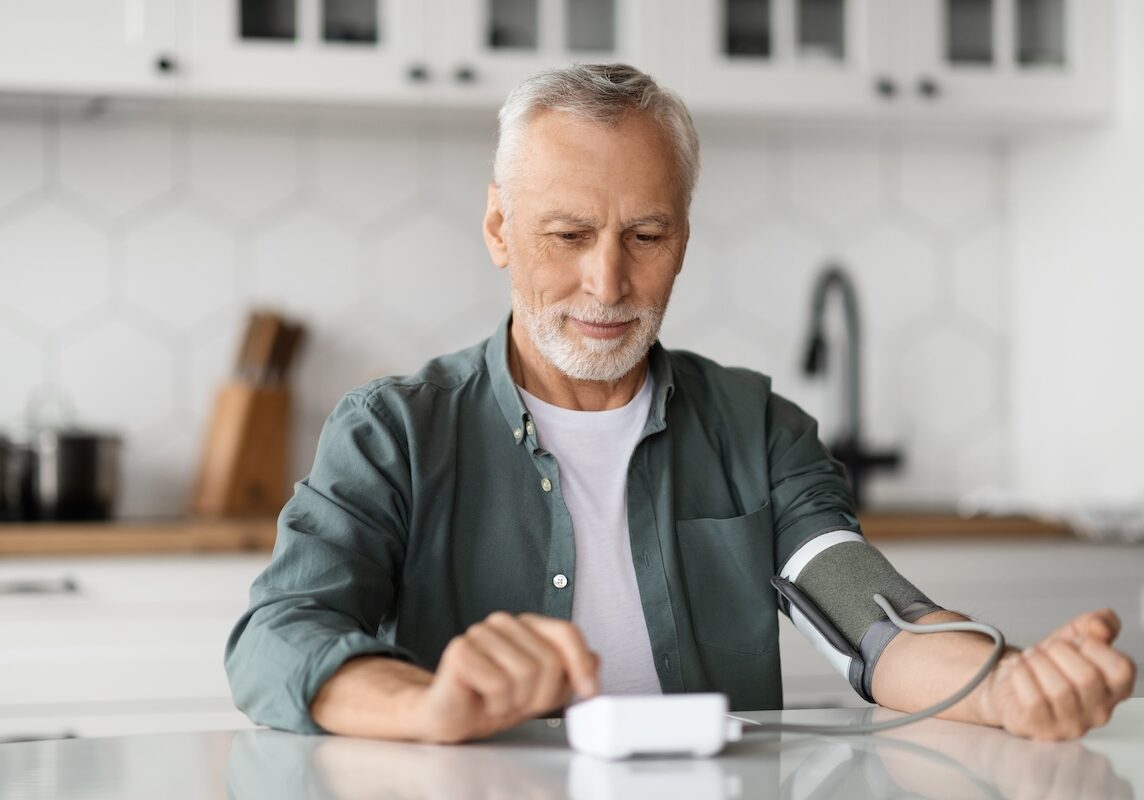 Senior Man Checking Blood Pressure With Upper Arm Monitor In Kitchen Interior, Smiling Elderly Gentleman Measuring Arterial Tension At Home, Controlling His Health Condition, Copy Space