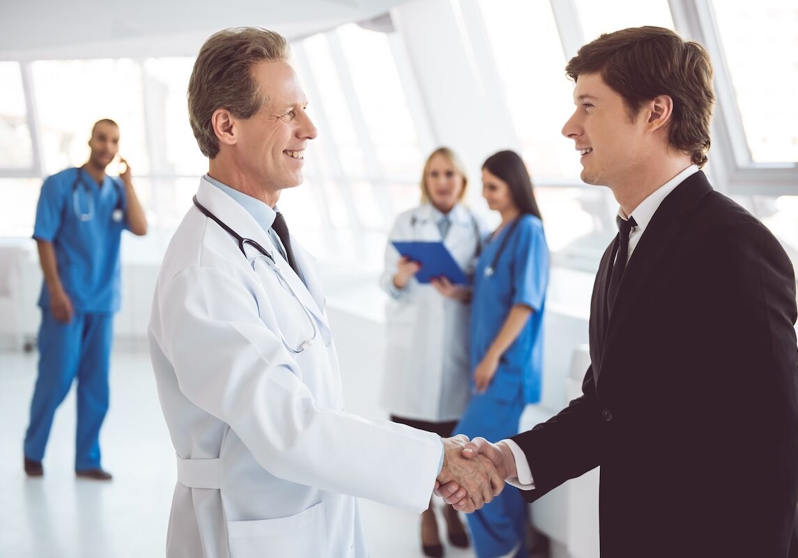 Handsome mature doctor and young businessman are shaking hands and smiling while standing in the hospital hall