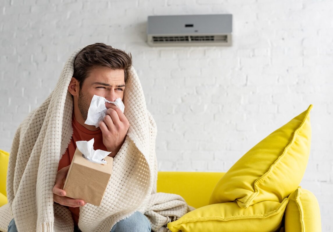 sick man with paper napkins warming under blanket while sitting under air conditioner at home